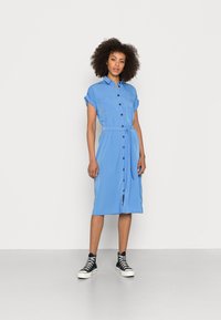 Robe chemise bleu clair avec des manches courtes, des boutons sur le devant, deux poches poitrine et une taille à nouer. Portée avec des sneakers montantes noires.