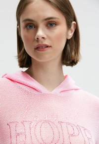 Junge Frau mit kurzen braunen Haaren trägt einen pinken, strukturierten Hoodie mit dekorativem Text auf der Vorderseite, vor einem schlichten, hellen Hintergrund.
