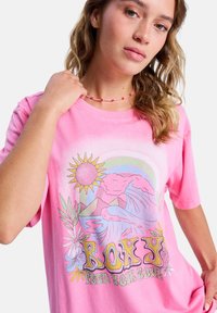 T-shirt en coton rose présentant un graphisme coloré avec un soleil, des montagnes et des vagues. Le design comprend des accents floraux et un col arrondi.