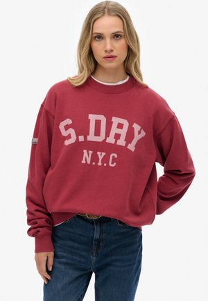 Woman with long blonde hair wears a red oversized sweatshirt with "S.DRY N.Y.C" text and blue jeans, posing against a white background.