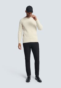 Romig geribbelde trui in combinatie met zwarte slim-fit broek en zwarte enkellaarsjes; een zwarte baseballcap maakt de look compleet.