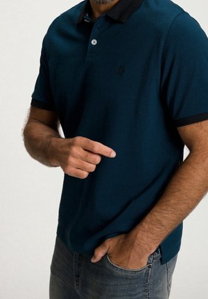 Man wearing a dark blue polo shirt with black collar and sleeve edges, one hand in jeans pocket, other hand pointing forward.