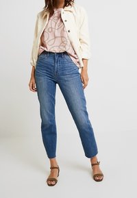 White cord jacket over a pink top with a gold pattern, paired with high-waisted blue jeans and brown ankle strap sandals.