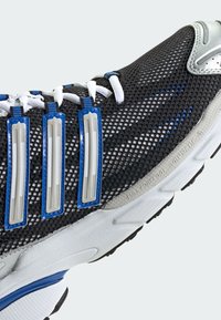 Scarpa sportiva nera e grigia con tomaia in mesh, dettagli blu e tre strisce grigie. Lacci bianchi e suola testurizzata per la presa.