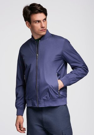 Jonge man die een marineblauw bomberjack met rits draagt, met zijn handen in de zakken, en naar links kijkt tegen een effen achtergrond.