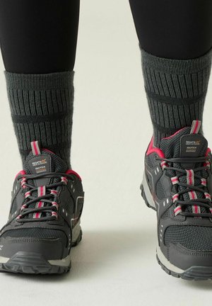 Scarponcini da trekking grigio scuro e rosa con spesse calze grigie a coste indossate da una persona che sta in piedi su uno sfondo neutro.