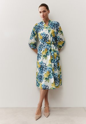 Frau trägt ein weißes Midikleid mit blauen Blumen- und gelben Zitronenmustern, dreiviertellangen Puffärmeln und beigefarbenen spitz zulaufenden Absätzen.