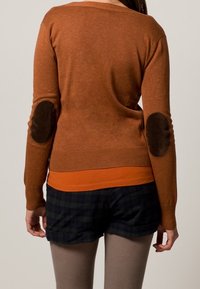 Sweter w kolorze rdzy z szerokim dekoltem i prążkowanym wykończeniem, z brązowymi zamszowymi łatami na łokciach. Połączony z pomarańczową koszulką pod spodem i szortami w kratę.
