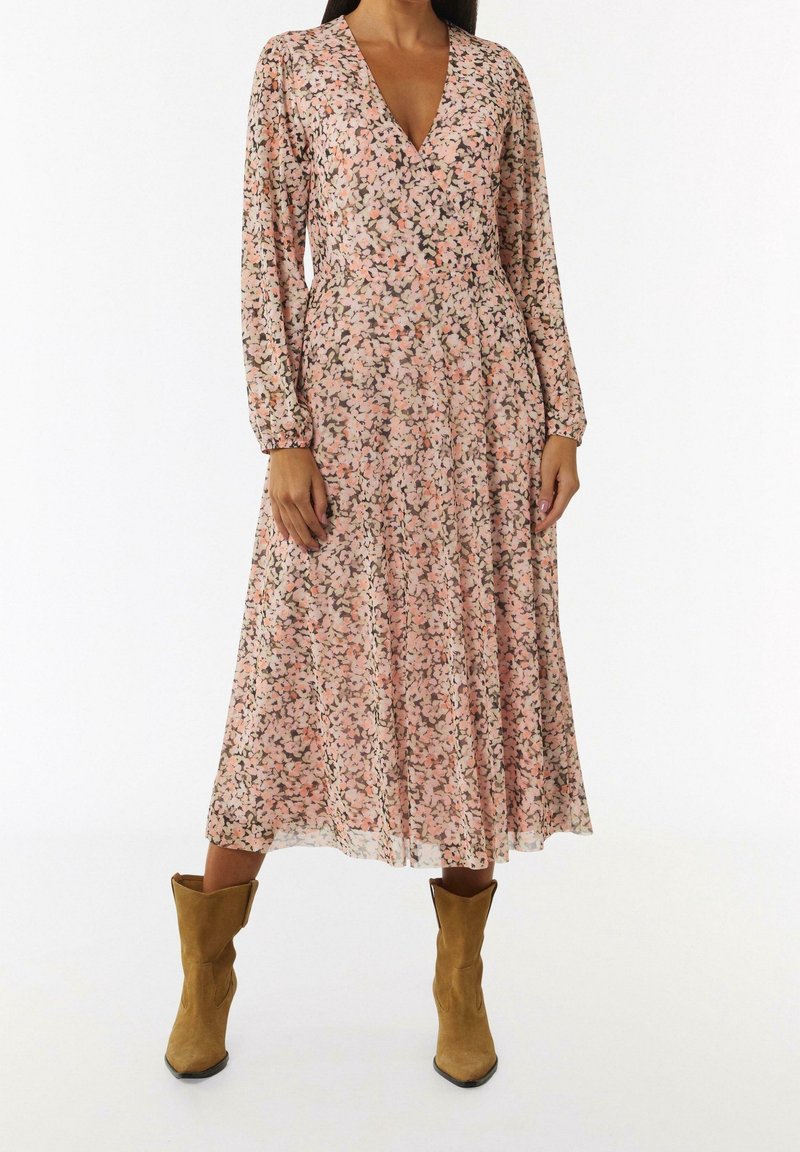 Femme portant une robe midi florale rose à manches longues avec un décolleté en V, associée à des bottes de cowboy en daim beige sur fond blanc.