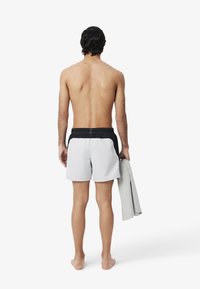 Zwartgrijze zwemshorts voor mannen met een elastische zwarte tailleband. De persoon houdt een grijze handdoek vast en heeft blote voeten. Het ontwerp is eenvoudig en functioneel.