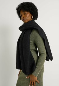 Bufanda negra con textura drapeada sobre un vestido de punto verde, que presenta un diseño holgado y oversized, con una apariencia suave y acogedora.