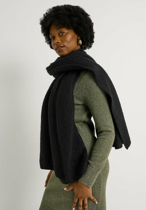 Bufanda negra con textura drapeada sobre un vestido de punto verde, que presenta un diseño holgado y oversized, con una apariencia suave y acogedora.