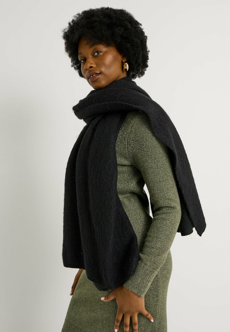 Bufanda negra con textura drapeada sobre un vestido de punto verde, que presenta un diseño holgado y oversized, con una apariencia suave y acogedora.