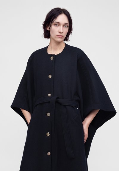 Γυναίκα με κοντά σκούρα μαλλιά, φοράει μαύρο παλτό με κουμπιά, φαρδιά μανίκια και ζώνη στη μέση, στέκεται μπροστά σε απλό ανοιχτό φόντο.