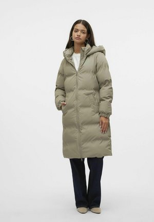 Manteau long et rembourré en vert olive atténué, avec capuche, fermeture éclair, poignets élastiques et poches latérales, porté sur un jean foncé.