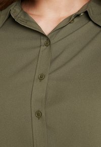 Chemise boutonnée vert olive avec col, texture lisse et six boutons assortis le long de la patte de boutonnage avant.