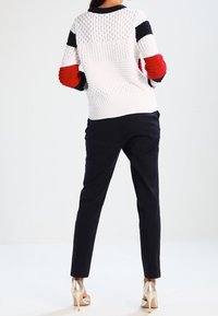 Vit stickad tröja med röda och marinblå detaljer, strukturerad design. Kombinerad med marinblå byxor och glänsande guldklackar, sedd bakifrån.