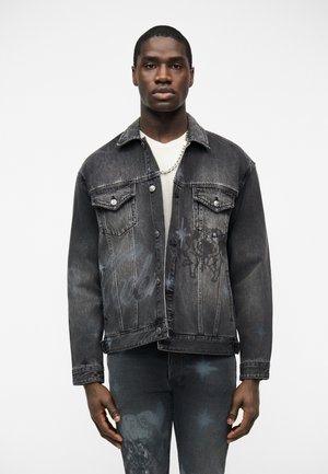 Homme portant une veste en denim noir délavé avec des poches avant et un jean assorti avec des imprimés subtils d'étoiles et graphiques, une chemise blanche et un collier en chaîne argenté.