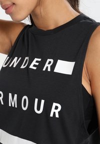 Detailní záběr na osobu v černém bezrukávovém topu s výrazným bílým textem "UNDER ARMOUR" přes hruď.