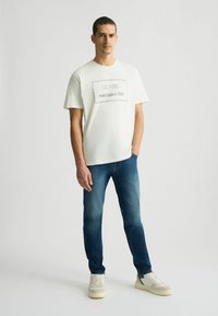T-shirt di cotone bianco con logo ricamato nero. Abbinato a jeans di denim blu. Modello in piedi con le mani rilassate, indossando sneakers beige.