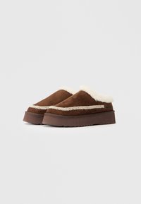 Brūni zamša slip-on apavi ar balto pūkaino oderi un dekoratīvām šuvēm. Bieza brūna gumijas zole ar strukturētu saķeres rakstu.