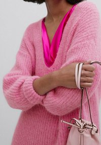 Różowy sweter z dzianiny w serek i teksturowanym wykończeniu, nałożony na błyszczący różowy satynowy top. Trzyma małą torebkę w kolorze kremowym.