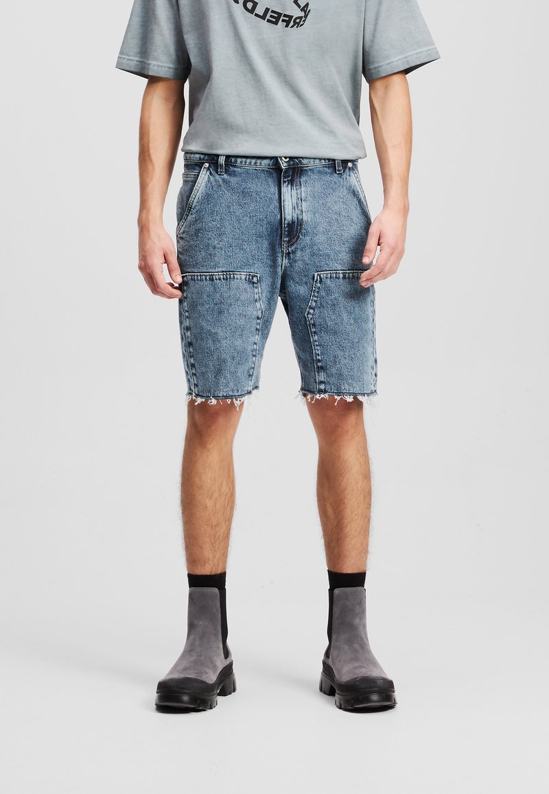 Shorts en denim bleu clair avec des bords effilochés, dotés de deux grandes poches avant, associés à un t-shirt oversized gris et des bottines grises.