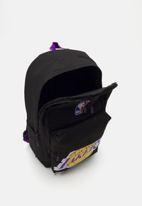 Sac à dos noir avec des accents violets, grande illustration sur le devant avec "Lakers", deux compartiments zippés et une boucle de suspension en haut. Tissu résistant.