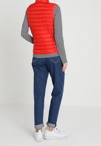 Röd pufferväst med vertikal söm, bärs över en svartvit randig långärmad tröja. Blå jeans med uppvikt kant och vita sneakers fulländar looken.