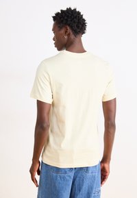Camiseta de algodón amarilla clara con mangas cortas, cuello redondo y un corte relajado, combinada con pantalones cortos de mezclilla azules. Vista trasera.