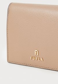 Portefeuille en cuir beige avec surface texturée, présentant un logo doré en relief "FURLA" et des coutures soignées le long des bords.