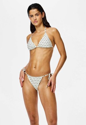 Femme aux cheveux mouillés portant un bikini blanc à pois noirs, debout devant un fond blanc uni.