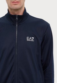 Jachetă bleumarin cu fermoar, realizată dintr-un material neted și ușor; prezintă un guler înalt și logo-ul EA7 pe piept. Design minimal și croială.