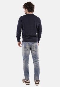 Felpa della marina, con polsini e orlo a costine, indossata con jeans blu sbiaditi. Sneakers con accenti marroni e bianchi, vista da dietro.