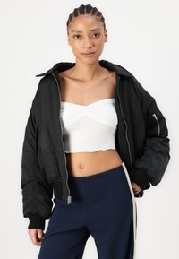 Bomber noir avec fermeture éclair et poignets côtelés, porté sur un haut court blanc. La tenue comprend un pantalon marine avec une bande sur le côté.
