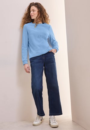 Ljusblå texturerad tröja med långa ärmar, kombinerad med mörkblå vida jeans och vita sneakers, mot en neutral bakgrund.