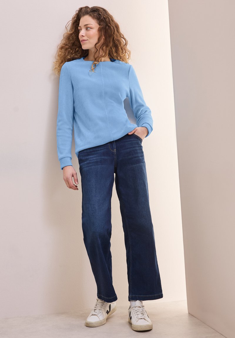 Hellblauer strukturierter Pullover mit langen Ärmeln, kombiniert mit dunkelblauen weiten Jeans und weißen Sneakers, vor einem neutralen Hintergrund.