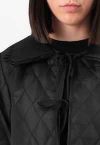 Femme portant une veste noire matelassée avec un grand col et des liens en ribbon sur un pull noir, coupé à la bouche et à la longueur des cheveux.