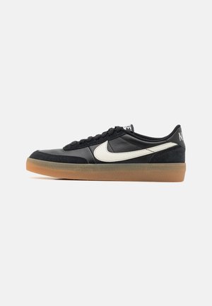 Zapatilla deportiva negra de corte bajo con el logo blanco de Nike y suela de goma color caramelo, vista lateral sobre fondo liso gris claro.