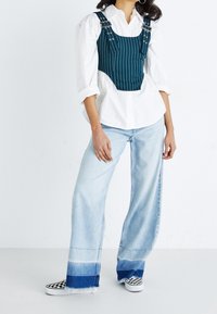 Sinisenvihreä raidallinen korsettitoppi, jossa säädettävät olkaimet, puettuna valkoisen paidan päälle, yhdistettynä vaaleansinisillä leveälahkeisilla farkuilla ja ruudullisilla slip-on-kengillä.