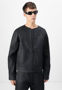Dark denim jacket features a zip front, round neckline, and quilted zigzag stitching. Model also wears sleek, angular black sunglasses.