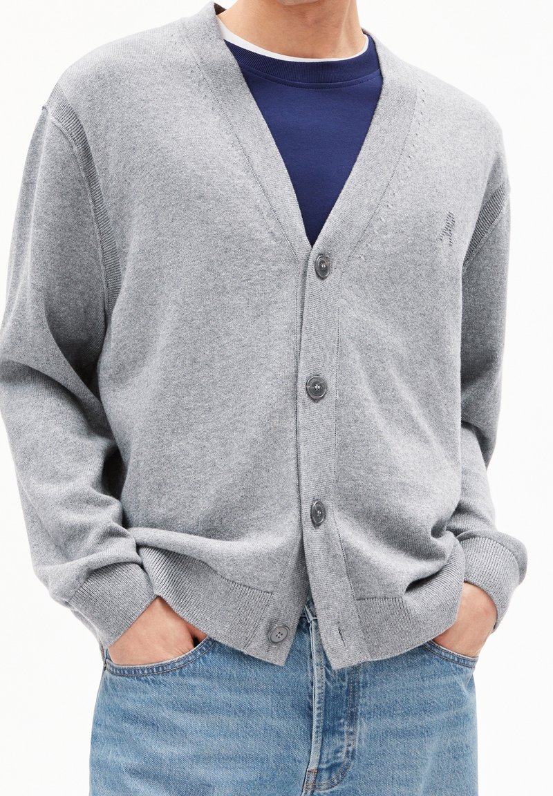 Homme portant un cardigan gris clair boutonné sur une chemise bleu marine, les mains dans les poches d'un jean bleu sur fond blanc.