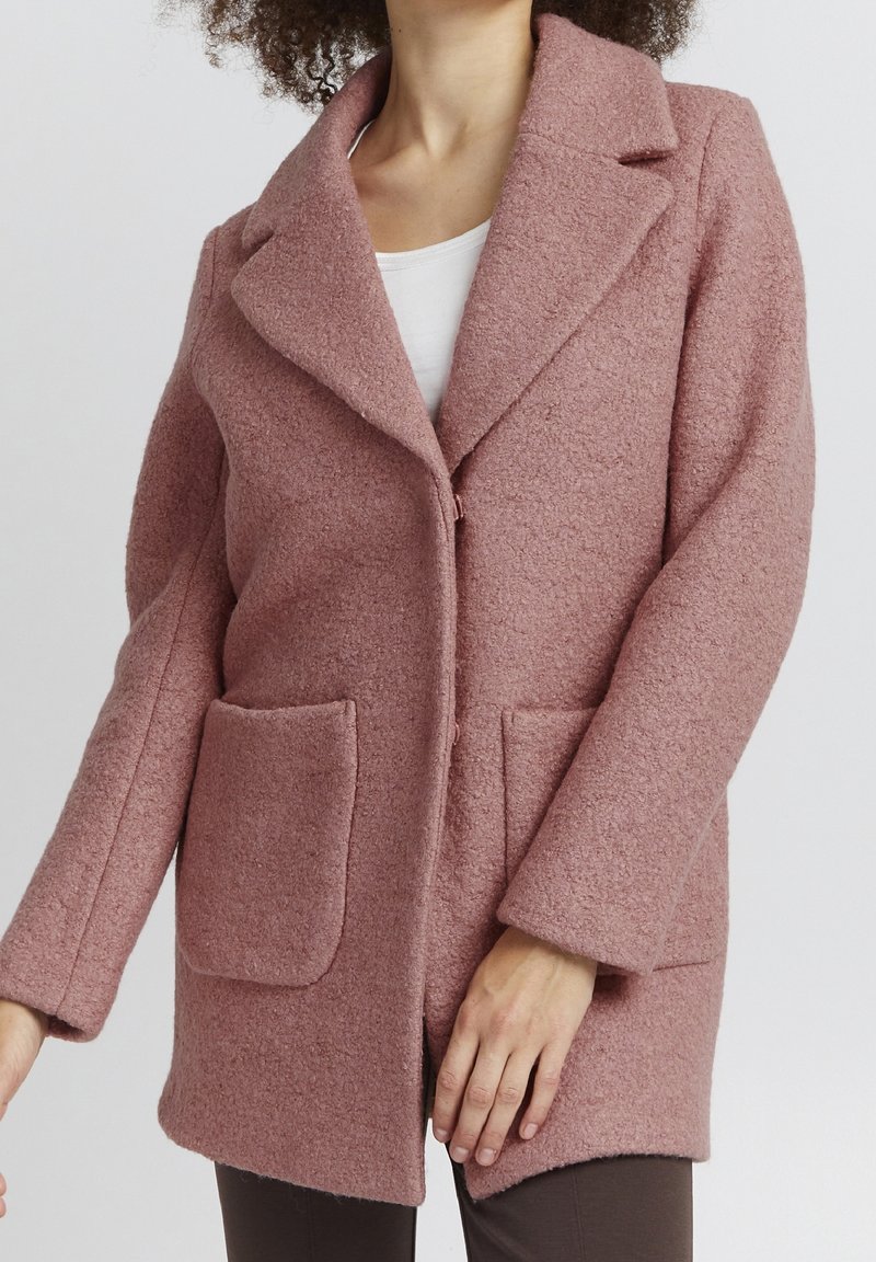 Femme portant un manteau en laine rose poudré texturé avec deux grandes poches avant, par-dessus un haut blanc, sur fond uni.