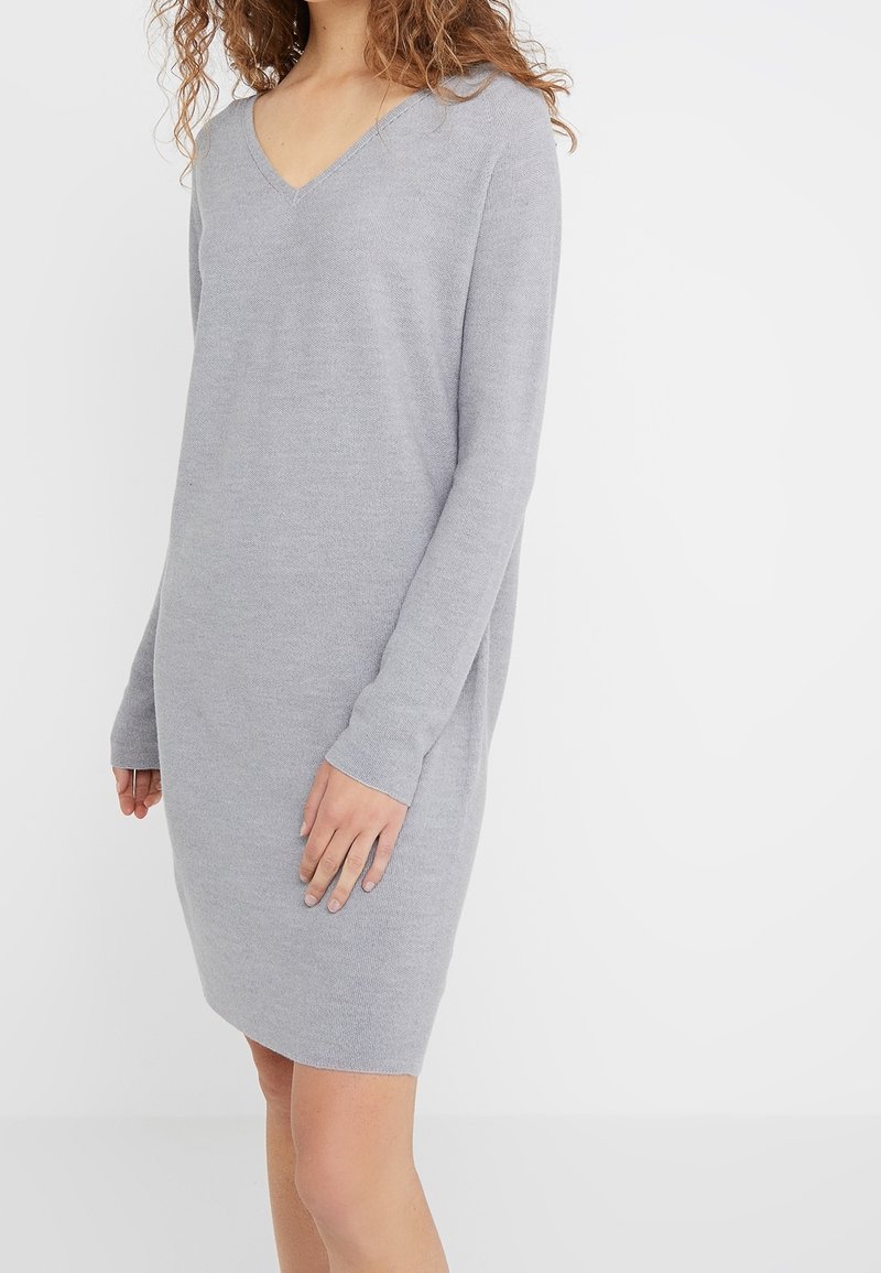 Robe tricotée grise avec un décolleté en V, des manches longues et une coupe décontractée. Le tissu a une texture lisse, arrive au genou sans motifs ni accents visibles.