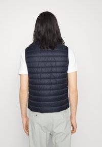 Chaleco acolchado azul marino con costuras horizontales, material ligero y sin mangas. Lleva puesto sobre una camisa blanca, con una textura suave.