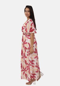 Femme dans une robe fluide à motif floral aux teintes rose et pêche, se tenant de côté, portant des sandales à talons hauts transparentes.