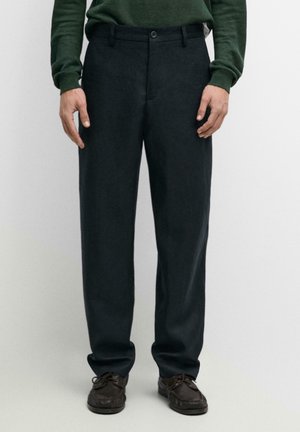 Mann trägt dunkelgraue maßgeschneiderte Hose, grünen Langarmpullover und braune Schnürschuhe, steht vor einfachem Hintergrund.