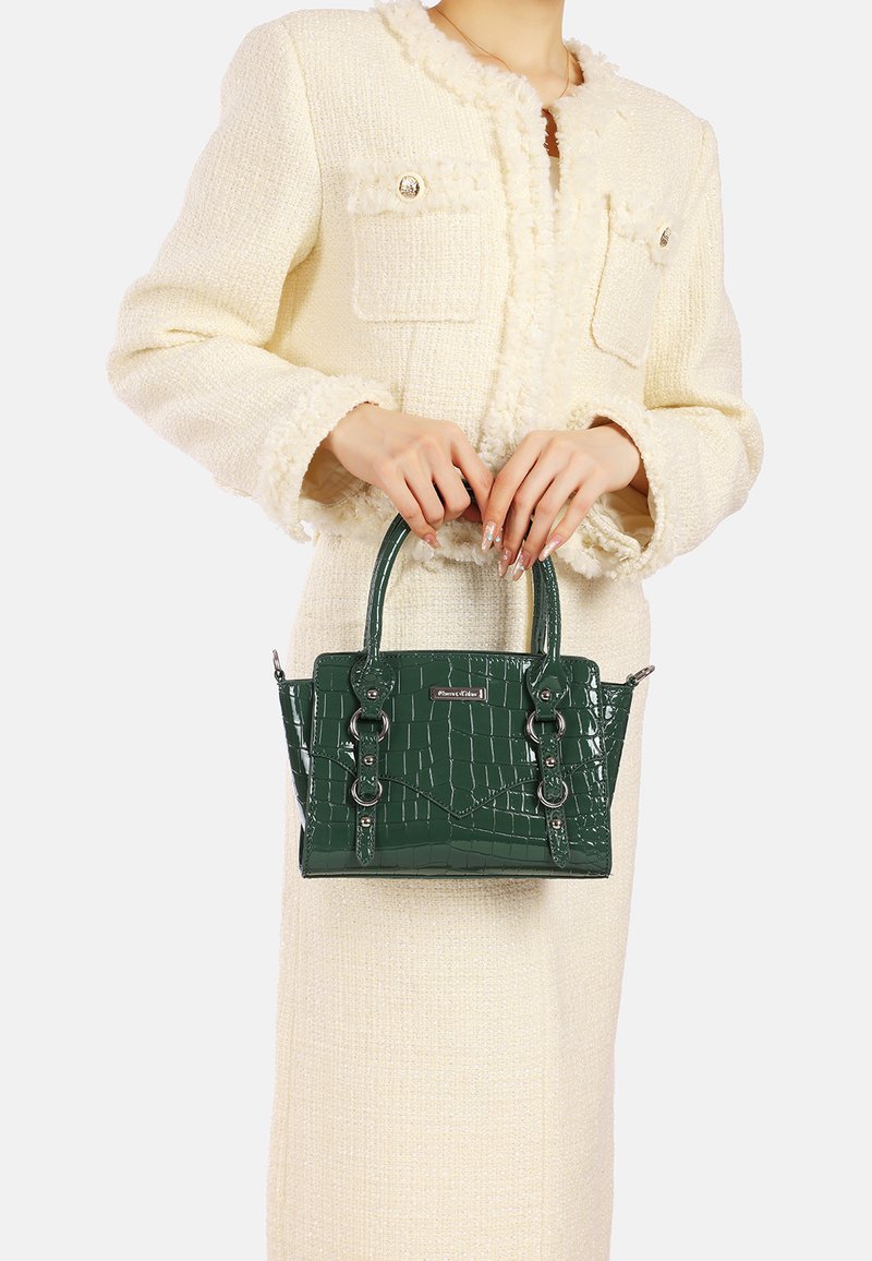 Donna in cappotto texturizzato color crema che tiene una borsa a tracolla di pelle con motivo coccodrillo verde scuro, dotata di manici doppi e rifiniture in metallo.