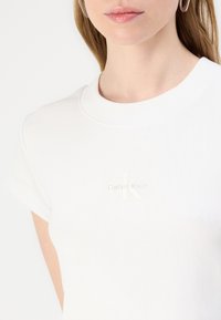 White ribbed T-shirt with short sleeves, featuring a subtle, embossed Calvin Klein logo on the chest. Smooth texture and classic fit.
