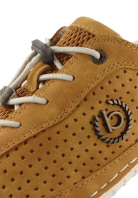 Bruna mockasiner i mocka med mesh-detaljer, perforerad textur, elastiska snören och en cirkulär logotyp på sidan. Sulan är lätt och flexibel.